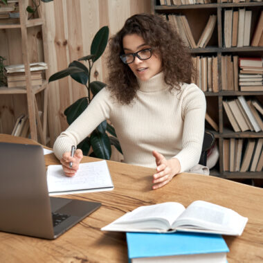 Mulher sentada em uma mesa, enquanto escreve e olha a tela do computador. Ao fundo, há uma estante de livros.