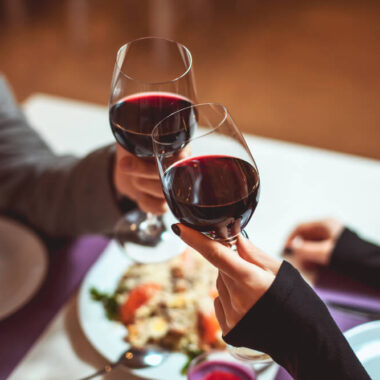 Casal fazendo um jantar com vinho tinto e brindando com as taças.