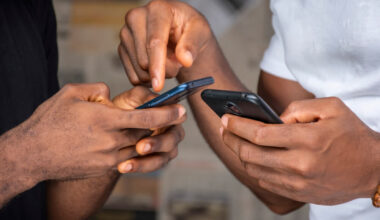 Close na mão de dois homens negros, cada um está segurando um aparelho celular diferente.