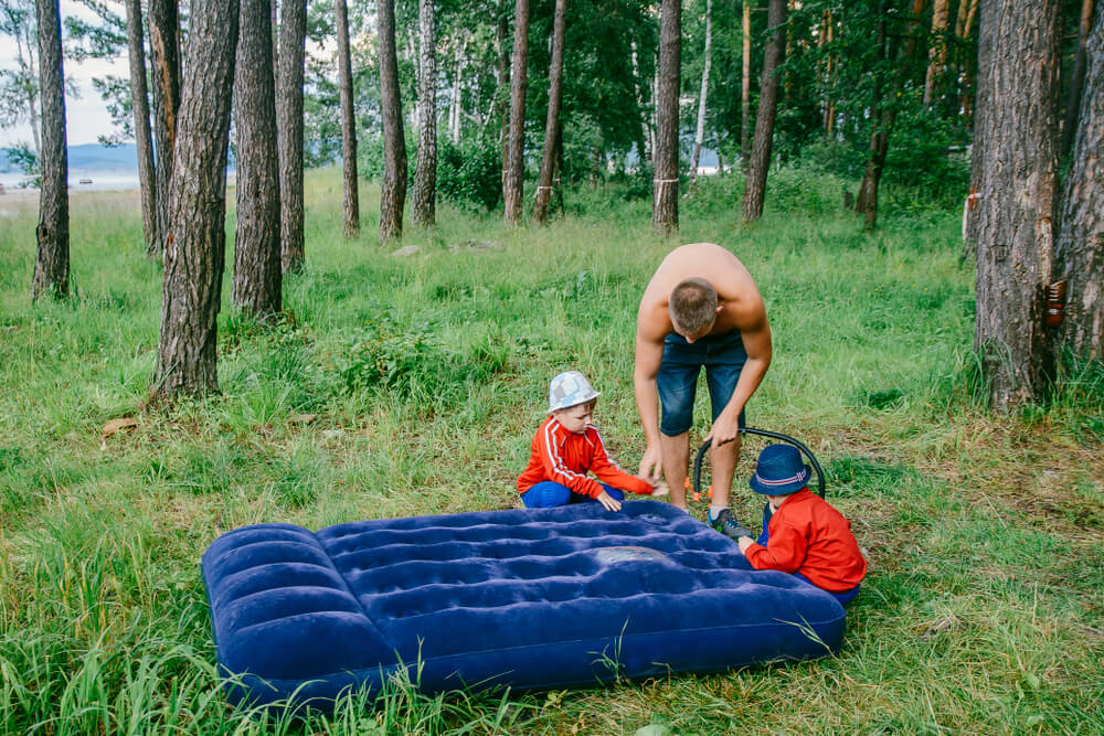 Homem e duas crianças em meio á uma floresta, enchendo um colchão inflável - um dos acessórios de camping.