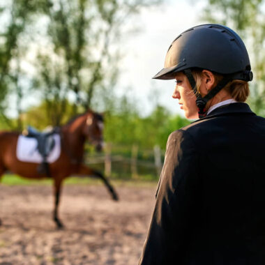 Mulher utilizando capacete para hipismo em primeiro plano. Em segundo plano, um cavalo.