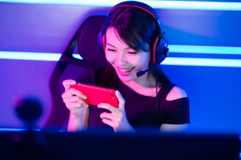 Mulher sentada em uma cadeira gamer, enquanto mexe no celular e está com um fones de ouvido gamers hyperx.