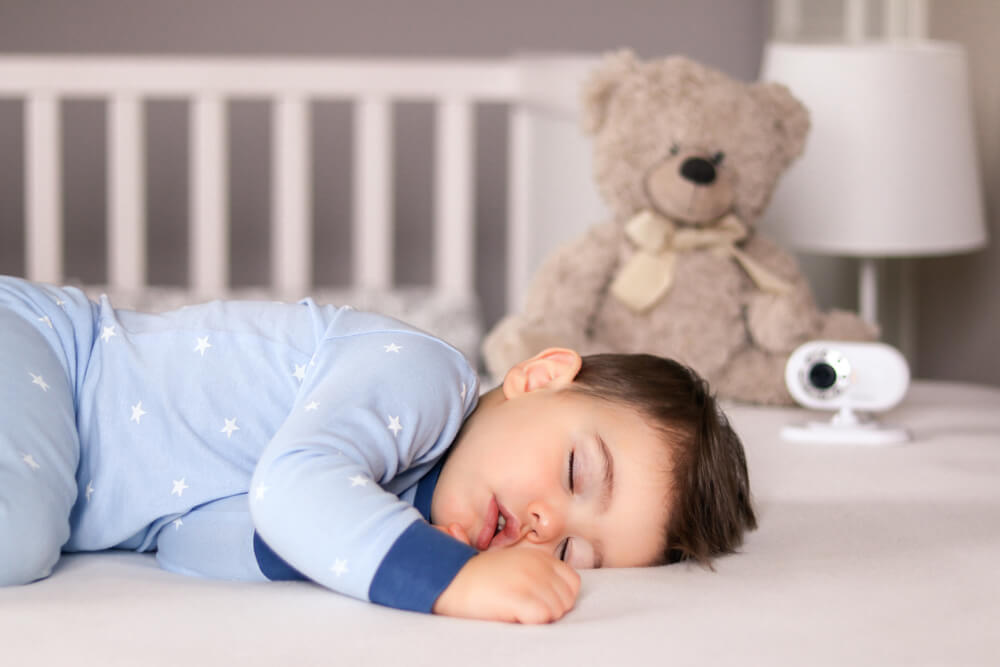 Bebê dormindo. Ao fundo da imagem, desfocados, vemos um urso de pelúcia e uma babá eletrônica monitorando o sono da criança.