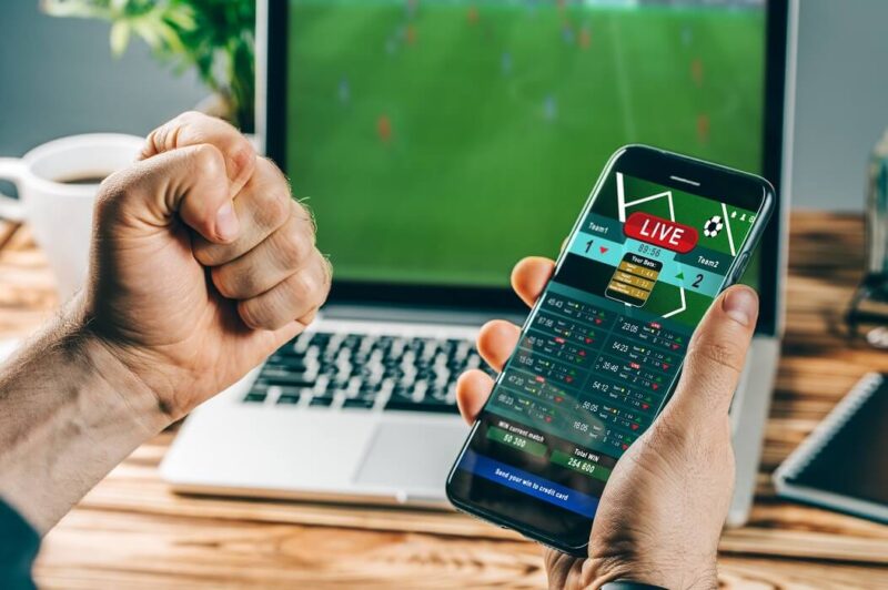 homem segurando o celular e acessando um dos melhores sites de apostas esportivas. Ao fundo, um notebook com um jogo de futebol na tela.